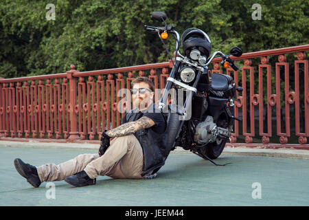 bearded redhead biker with beard in leather jacket sitting on floor near bike. Stock Photo