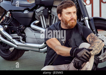 bearded redhead biker with beard in leather jacket sitting on floor near bike. Stock Photo