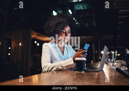 Young businesswoman working at late night shift and using mobile phone. African female looking at her smartphone while working. Stock Photo
