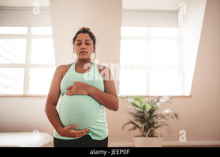 Portrait of young pregnant woman standing at home with her eyes closed and holding belly. pregnant female meditating at home. Stock Photo