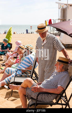 Dickensian style beach party, Row of men sitting, with one standing, in deckchairs all wearing straw hats and Victorian bathing costumes. Stock Photo