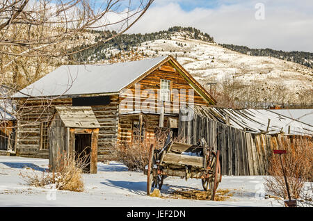 Log home, outhouse, and a wagon on a winter day in a ghost town with a bit of blue sky and sunlight Stock Photo