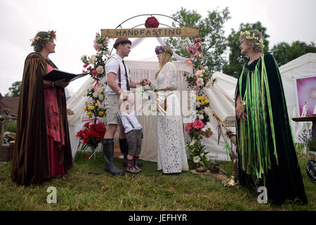 Fi Sexton, 32 and Peter Howe, 38, both from London accompanied by son Thomas Howe, 2 take part in a Handfasting wedding ceremony. The ceremony performed by Glenda proctor (brown) and Jane Tove (green) involved tying their hands together with bonds, Jumping a broom and Eating cake and drinking ale together at Glastonbury Festival, at Worthy Farm in Somerset. Stock Photo