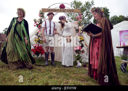 Fi Sexton, 32 and Peter Howe, 38, both from London accompanied by son Thomas Howe, 2 take part in a Handfasting wedding ceremony. The ceremony performed by Glenda proctor (brown) and Jane Tove (green) involved tying their hands together with bonds, Jumping a broom and Eating cake and drinking ale together at Glastonbury Festival, at Worthy Farm in Somerset. Stock Photo