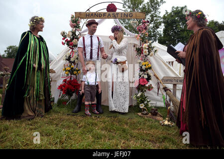 Fi Sexton, 32 and Peter Howe, 38, both from London accompanied by son Thomas Howe, 2 take part in a Handfasting wedding ceremony. The ceremony performed by Glenda proctor (brown) and Jane Tove (green) involved tying their hands together with bonds, Jumping a broom and Eating cake and drinking ale together at Glastonbury Festival, at Worthy Farm in Somerset. Stock Photo