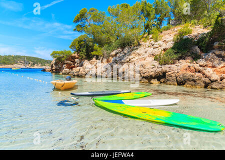 Yellow boat and surfing boards on idyllic Cala Portinatx beach and cliff rocks with pine trees in background, Ibiza island, Spain Stock Photo