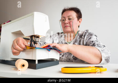 needlework and quilting - tailor sews on the sewing machine multicolored patchwork fabric sitting at a white table, on which are threads, buttons and rotary fabric cutters on white background. Stock Photo