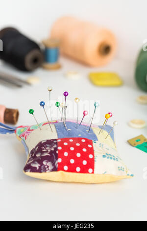 needlework, craft, sewing and tailoring concept - close-up on white desk with pincushion and pins, measuring meter, pink, blue and black thread spools, white buttons, soap, selective focus, vertical. Stock Photo