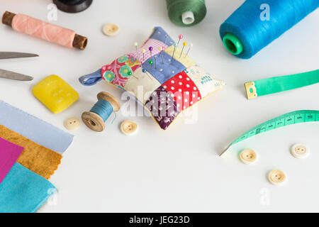 needlework, craft, sewing and tailoring concept - tools closeup on white desk, measuring meter, thread spools, pieces of colorful patchwork fabric, pincushion, buttons, soap, scissors. Stock Photo