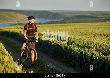 Attractive cyclist stands on the bike on the trail of the hay field in summer season. Stock Photo