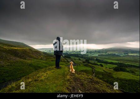 A man stands with his dog on a grassy hill looking out over the lush, green landscape under a stormy sky; North Yorkshire, England Stock Photo