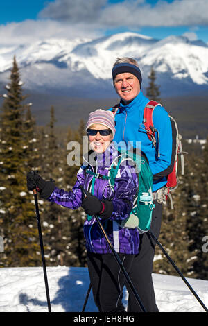 Male and female cross country skiiers on a lookout with snow covered mountains, blue sky and clouds in the background; Lake Louise, Alberta, Canada Stock Photo