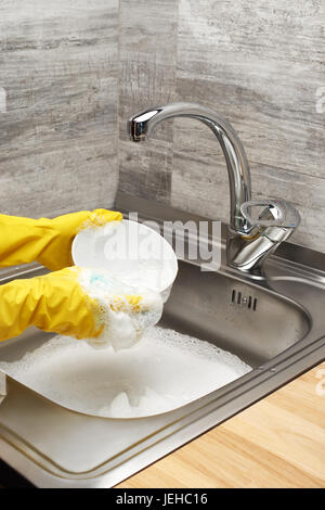 Close up of female hands in yellow protective rubber gloves washing white bowl with blue cleaning sponge against kitchen sink full of foam Stock Photo