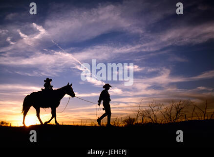 Agriculture - A cowboy leads his young daughter on a horse at sundown while carrying a fishing pole / Childress, Texas, USA. Stock Photo