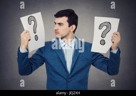 Perplexed young student businessman holding to white papers with question marks on them knowing what to do.Stressed young man having to many questions Stock Photo