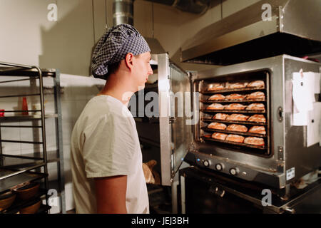 Handsome baker in uniform holding tray full of freshly baked bread at the manufacturing Stock Photo