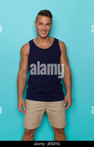 Handsome young man in blue tank top and beige shorts, standing relaxed, looking at camera and smiling. Three quarter length studio shot on turquoise b Stock Photo