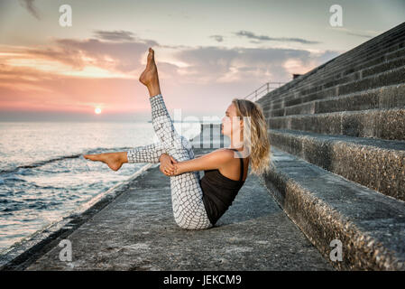 woman practicing yoga on concrete steps by the sea at sunset Stock Photo