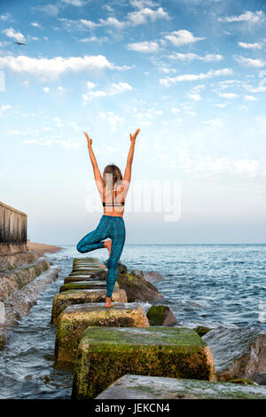 woman practicing yoga by the sea standing on rocks Stock Photo