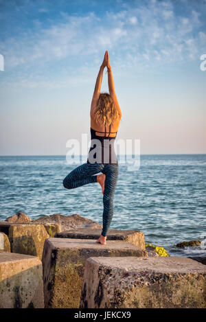 woman practicing yoga by the sea standing on rocks Stock Photo
