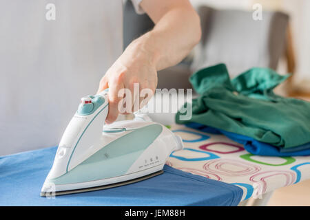 An iron on an ironing board with a blue shirt close up from the side with a stack of laundry Stock Photo