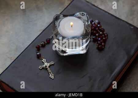 Close up of a weathered leather covered bible with a set of red rosary beads and a burning tealight candle in a glass candle holder. Stock Photo