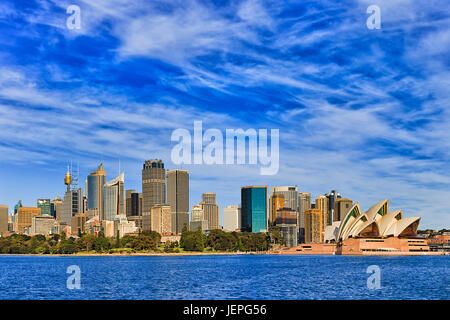 Cityscape of Sydney CBD across Royal Botanic gardens as seen from Sydney harbour ferry with blue water and sky. High-rise towers lit by bright warm su Stock Photo