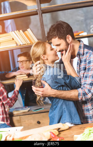 young people partying at home. Loving couple kissing while others conversing near bookshelf Stock Photo