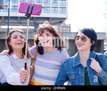 Three female friends taking a selfie in the city. They are using a smartphone on a selfie stick. Stock Photo