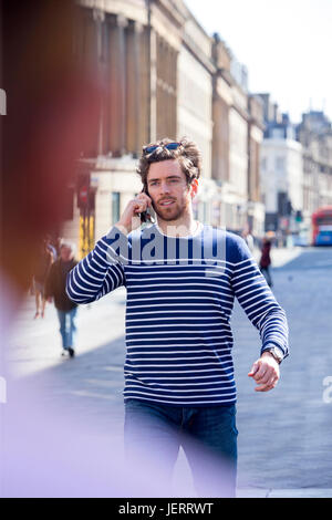 Man on the phone in the City. He is walking down the highstreet with his sunglasses on his head. Stock Photo