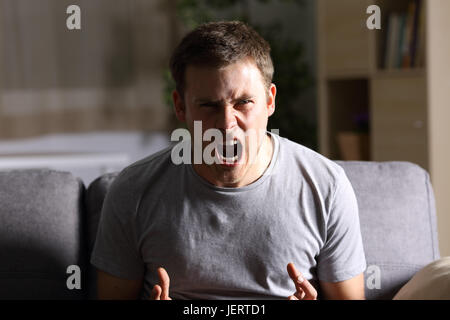 Casual furious man shouting and looking at you sitting on a sofa at home with a dark light in the background Stock Photo