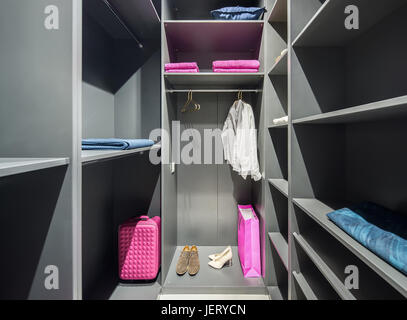 Illuminated gray walk-in closet with many shelves. There are shoes, shirts, hangers, towels, blue pillow, pink bags. Indoors. Horizontal. Stock Photo