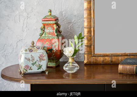Fancy colorful jug and casket with flower patterns on the wooden tabletop on the textured light wall background. Next to them there is a flower in a v Stock Photo