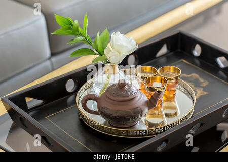 Metal plate on the dark wooden tray on the glass table on the background of the gray sofa. There is a clay teapot, three small glasses and a flower in Stock Photo