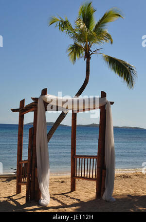 Wedding Arbor draped in white fabric located on the beach with a palm tree behind it. Stock Photo