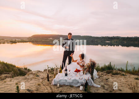 Thehappy newlyweds are holding hands during their picnic near the river during the sunset. Stock Photo