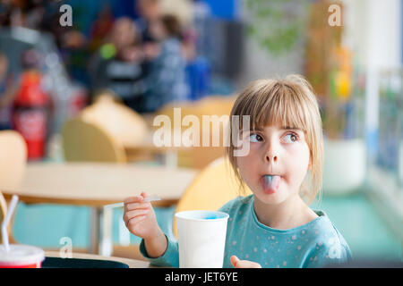 Cute blonde girl sticking or poking tongue out with blue crushed ice drink in brightly lit natural light cafe or restaurant Stock Photo