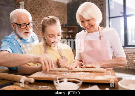 grandmother, grandfather and granddaughter cooking and kneading dough for cookies at kitchen table, cooking in kitchen concept Stock Photo