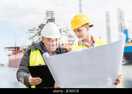Shipbuilding engineer discusses documents with a student. Experienced worker wearing white safety helmet explains technical issues to a manual laboure Stock Photo