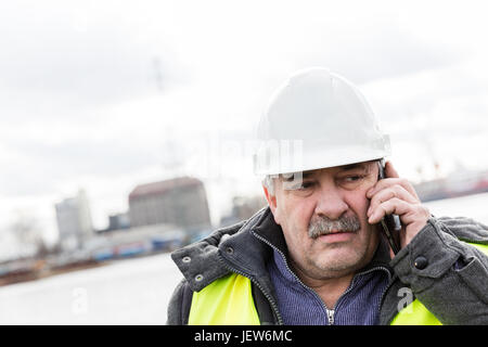 Senior engineer builder talking on the phone at the construction site in a port. Wearing safety helmet and yellow vest. Stock Photo