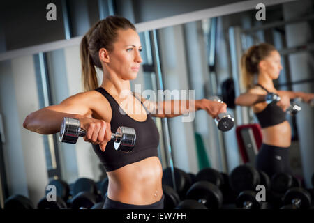 Young woman lifting dumbbells next to a mirror at a modern gym. Body training, weightlifting workout. Crossfit exercise. Stock Photo