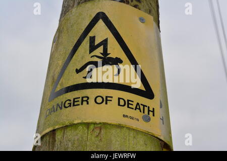 weathered yellow and black plastic danger of death sign tacked nailed onto wooden timber electricity pole pylon England UK Stock Photo