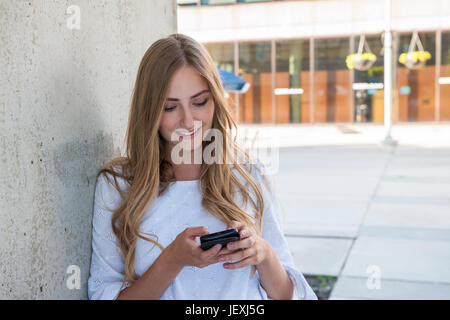 Young female university student on college campus using her smartphone and smiling standing outside near wall with copy space Stock Photo