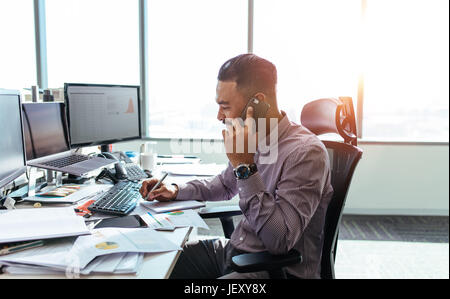 businessman talking on mobile phone and making notes. Man sitting at his office desk working on graphs and charts and discussing on phone. Stock Photo