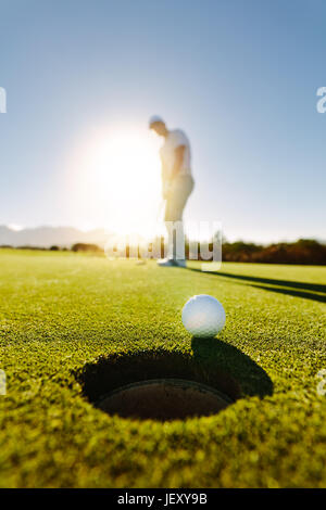 Vertical shot of professional golfer putting golf ball in to the hole. Golf ball by the hole with player in background on a sunny day. Stock Photo