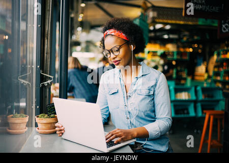 Focused young African woman sitting alone at a counter in a cafe working on a laptop and listening to music on earphones Stock Photo