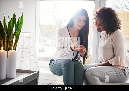 Two young woman seated on a windowsill with sun flare reading an sms or text message on a mobile phone with quiet smiles Stock Photo