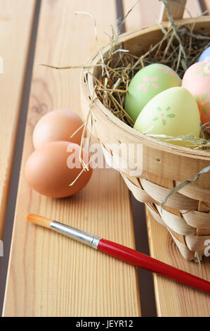 Hand painted Easter eggs in a basket filled with straw with natural, unpainted eggs and paintbrush resting on a wood plank table top. Vertical orienta Stock Photo