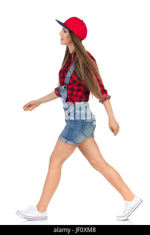 Woman in red lumberjack shirt, jeans shorts and white sneakers walking and looking away. Side view. Full length studio shot isolated on white. Stock Photo