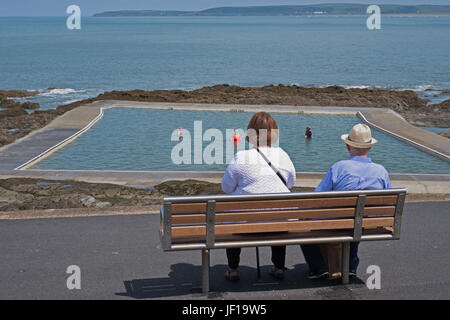 Two senior citizens watch bathers in a nineteenth century tidal rock pool on a hot summer day in Devon UK Stock Photo